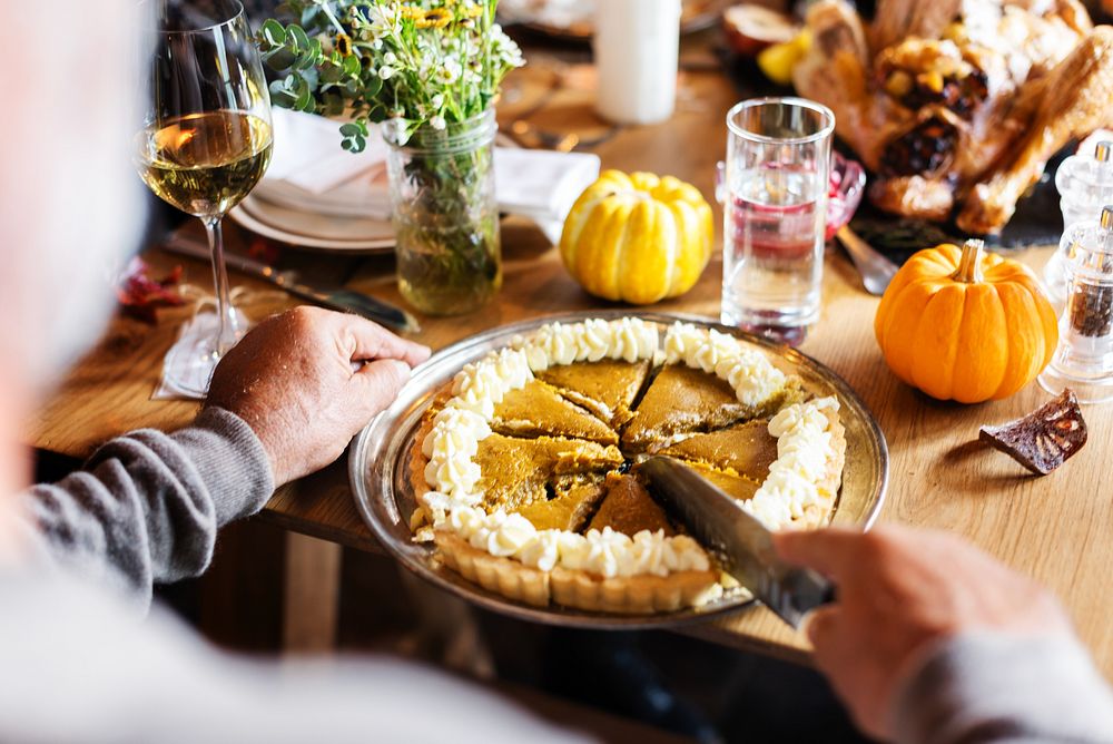 Pumpkin pie being sliced on a table with decorative pumpkins in Thanksgiving dinner. Warm autumn themed setting with people…