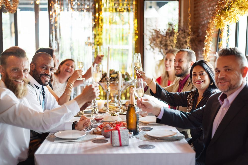 Group of diverse people celebrating at a festive dinner table, raising glasses in a toast, with decorations and a gift box…