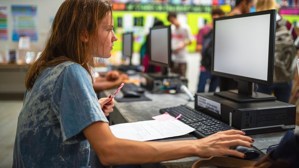 Person working at a computer in a busy office environment, focused on tasks. Office setting with computers and paperwork…