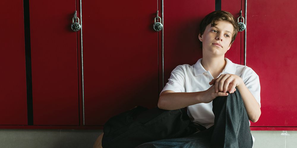 Teen student sitting by red lockers, wearing casual clothes. Student boy sitting depressed on school corridor. School boy…