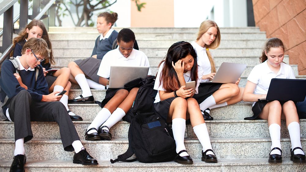 Students in uniforms sit on steps, using laptops and phones. Diverse group of boys and girls engaged in technology, studying…