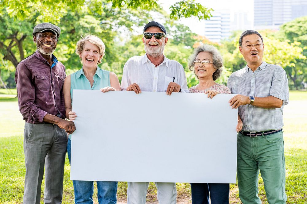 Diverse group of older adults holding a blank sign outdoors. Smiling seniors, men and women, enjoying a sunny day in the…