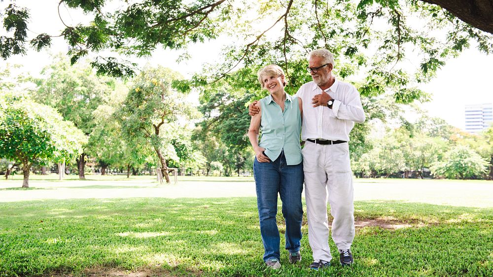 Elderly couple walking in a park, smiling and embracing. The elderly couple enjoys a sunny day, surrounded by trees, sharing…