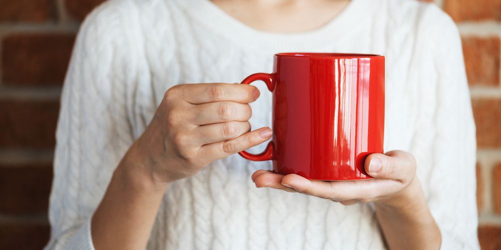 Person holding a large red mug, wearing a white sweater. Close-up of hands with a red mug. Red mug and sweater create a…