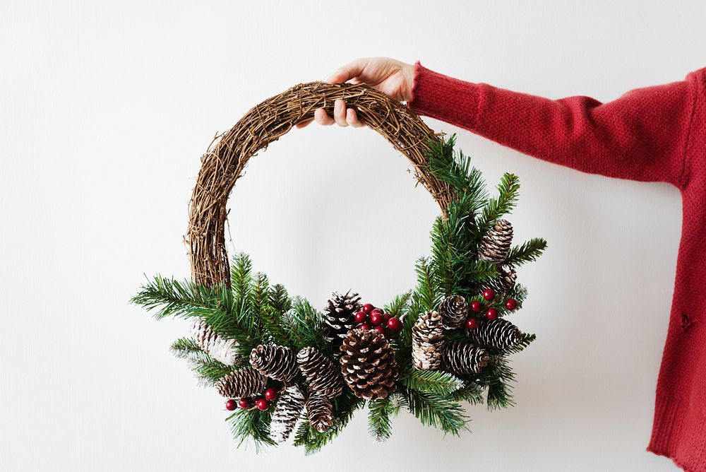 Hand holding a festive wreath with pinecones and greenery. Wreath features pinecones, greenery, and berries. Holiday wreath…