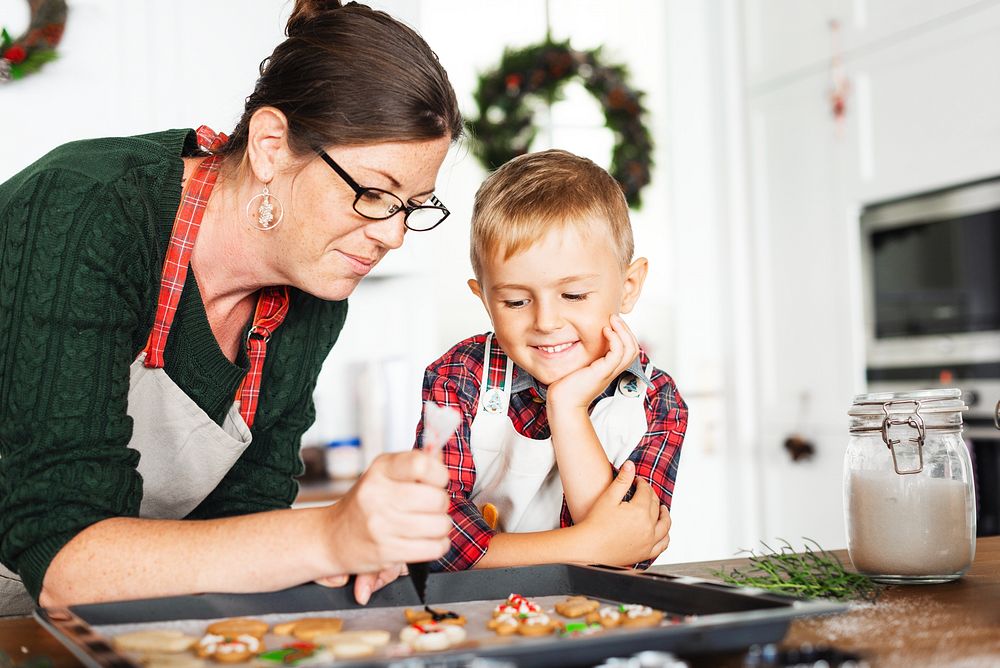 A woman and child decorate cookies in a kitchen. The woman wears glasses and an apron. The child watches eagerly. Holiday…