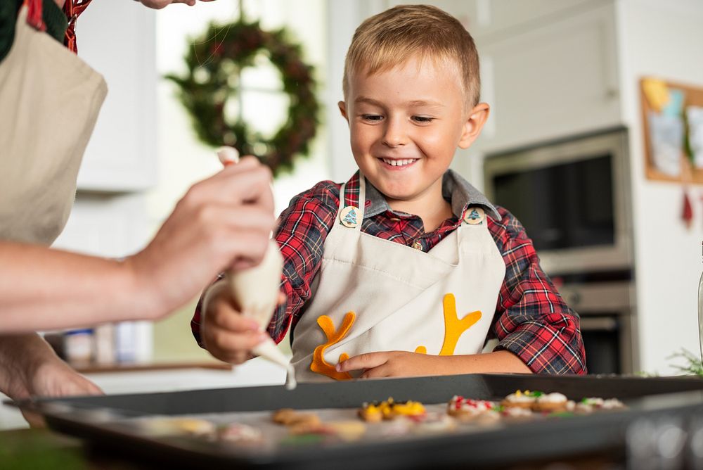 A cheerful young Caucasian boy in a plaid shirt enjoys baking cookies with a caregiver. The boy smiles as they decorate…