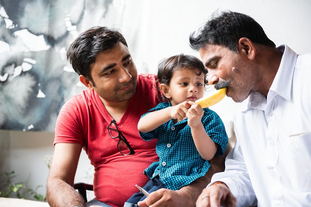 A Indian child feeds a man a slice of fruit while another Indian man watches. The scene captures a moment of care and…