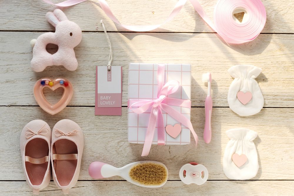 Flat lay of baby girl items: pink shoes, plush toy, gift box, and accessories. Soft pink and white theme for baby girl…