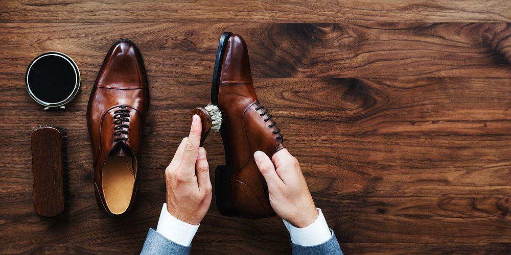 Hands polishing brown leather shoes on wooden floor. Shoe care with brush and polish. Elegant brown shoes, shoe care…