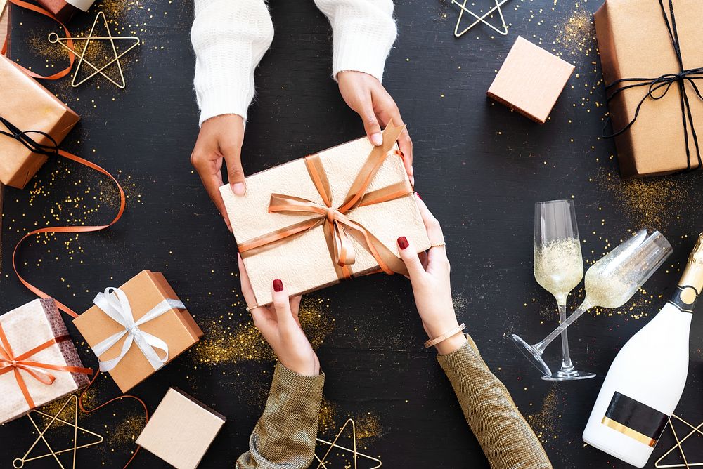 Hands exchanging a gift box with ribbon, surrounded by more gift boxes, champagne glasses, and festive decorations on a…