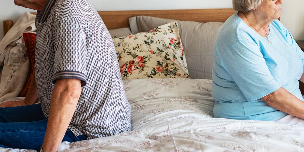 Elderly couple sitting back to back on bed, showing tension. The couple appears distant, highlighting relationship strain.…