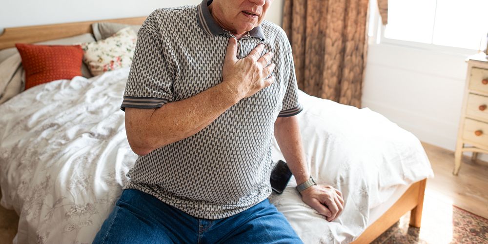 Elderly man sitting on bed, hand on chest, appearing unwell. Casual attire, bedroom setting. Concerned expression, elderly…