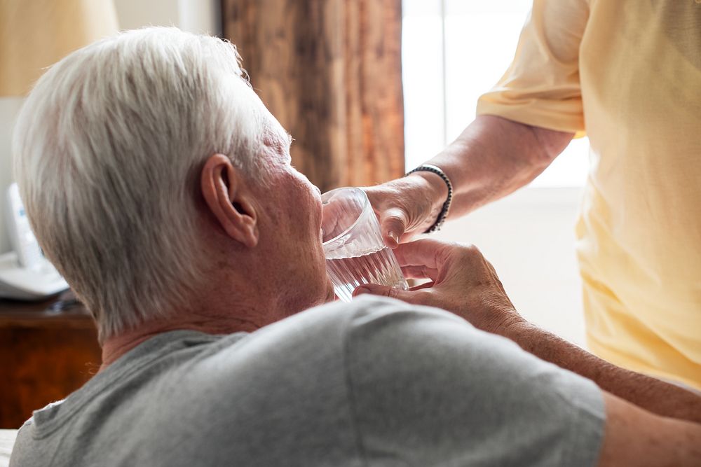 Elderly man drinking water with assistance. Senior care and support. Elderly nursing care and assistance. Woman taking care…