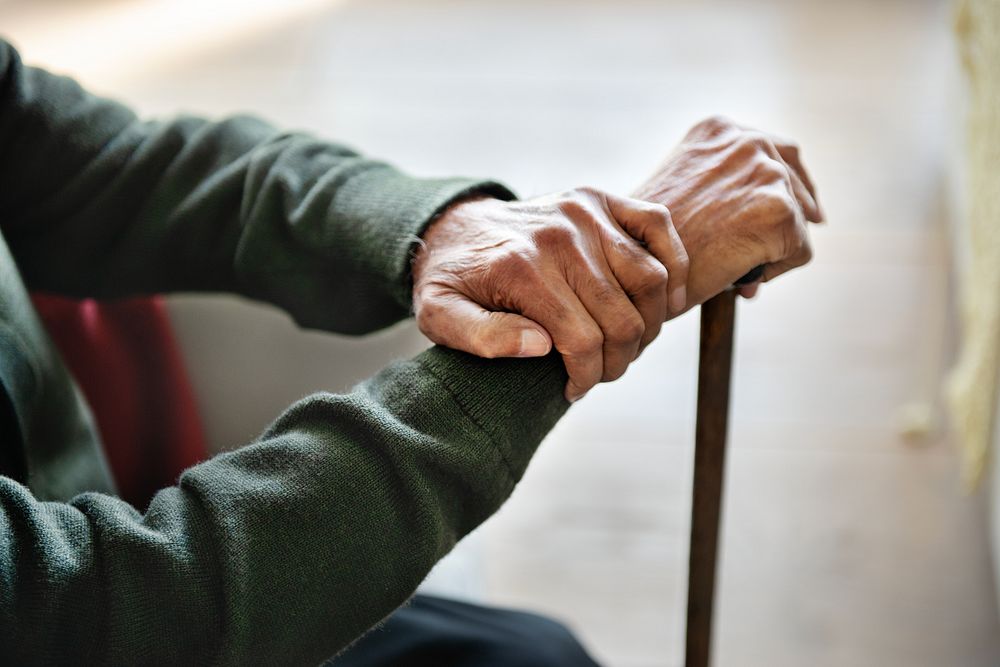 Close-up of elderly hands resting on a cane, symbolizing aging and support. The hands show wrinkles and veins, emphasizing…