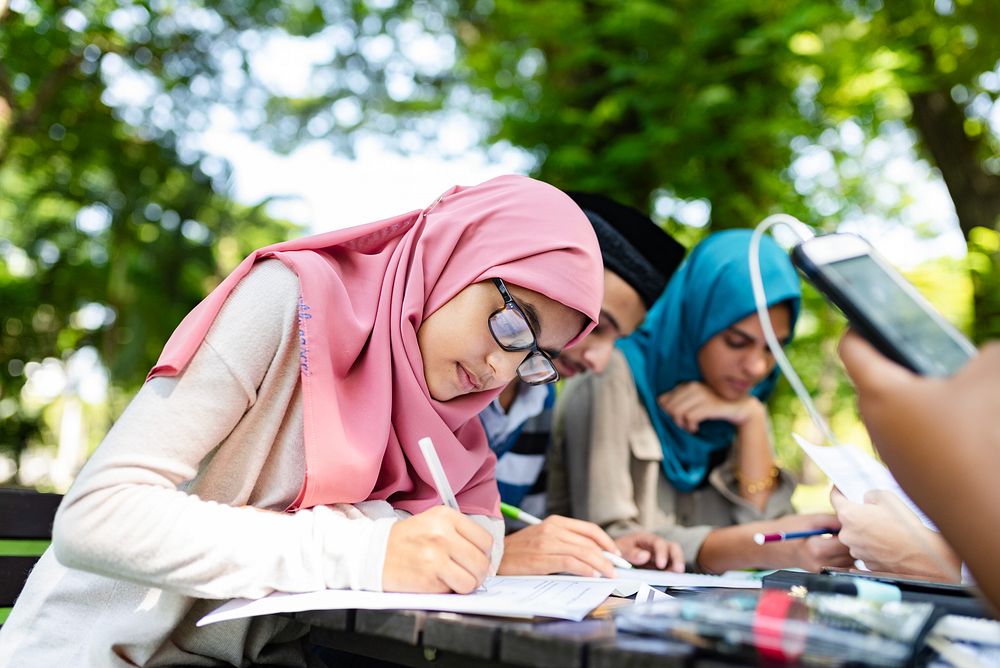 Group of students studying outdoors. Focused young woman in hijab writing. Diverse group engaged in learning. Education and…