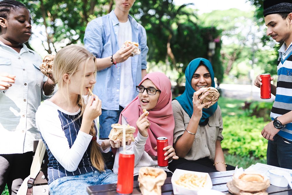 Group of diverse friends enjoying a picnic outdoors, eating sandwiches and drinking soda. Multicultural gathering with food…