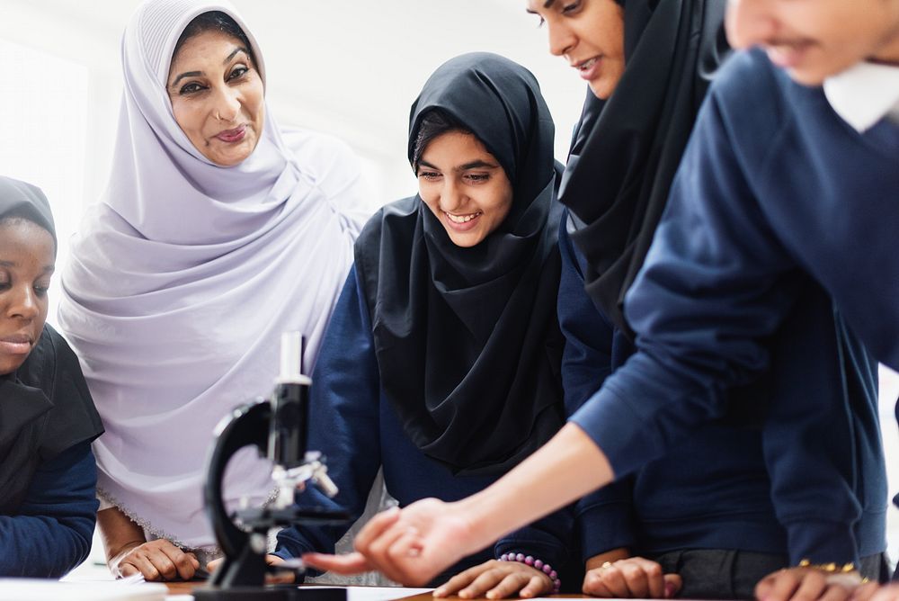 Group of students and a teacher in hijabs and uniforms, gathered around a microscope, engaged in a science lesson…