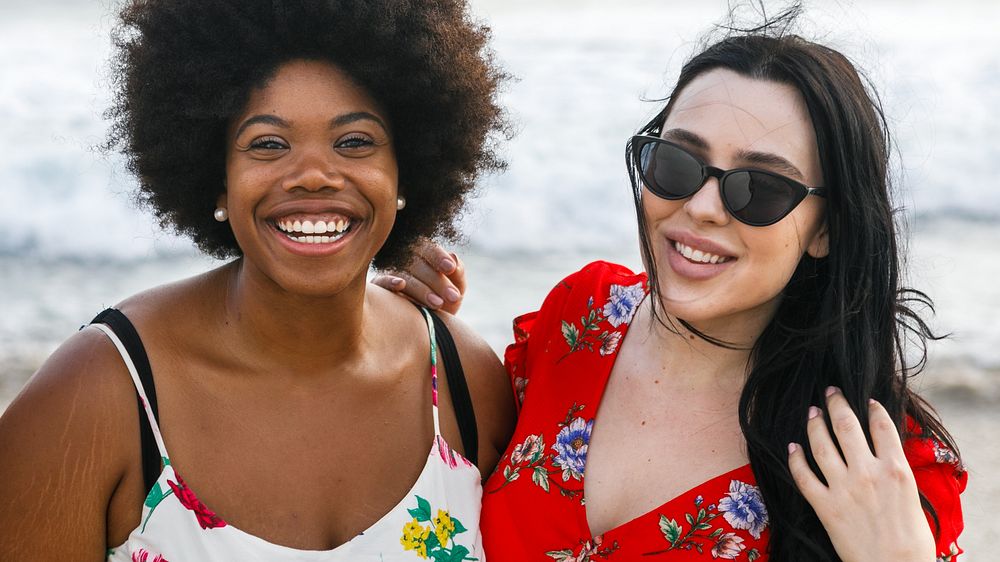 Diverse two women enjoying a beach day. African American woman wears a floral dress, the other a red floral dress. Both are…