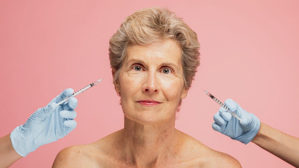 Elderly woman with short hair against a pink background. Two gloved hands hold syringes near her face, suggesting cosmetic…