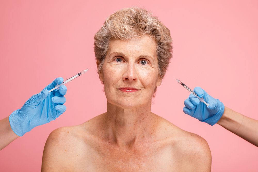 Senior woman with short hair, receiving cosmetic injections. Two hands with gloves hold syringes. Focused on beauty and…