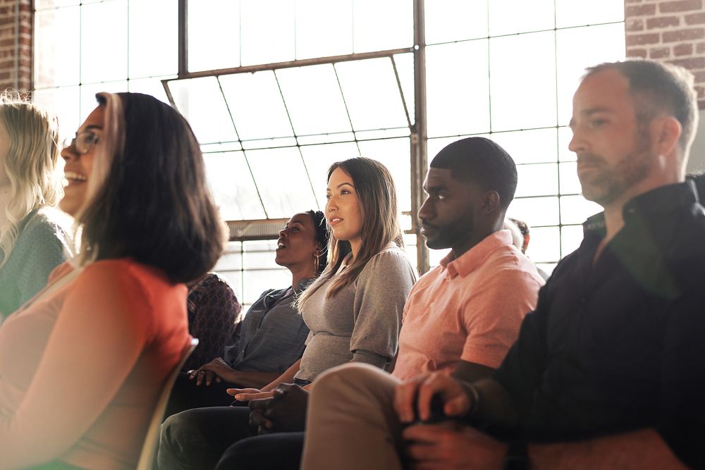 Diverse group of people sitting in a sunlit room, attentively listening. Mixed genders and ethnicities, focused and engaged…