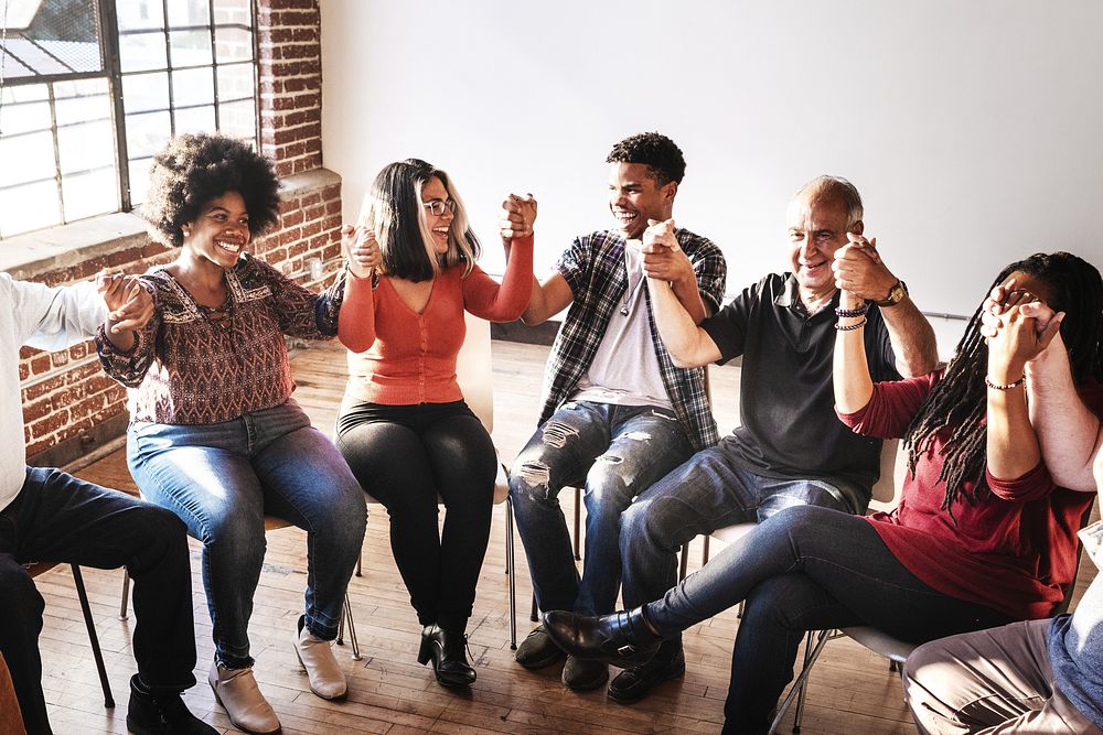 Diverse group of people sitting in a circle, holding hands, and smiling. Mixed genders and ethnicities, showing unity and…