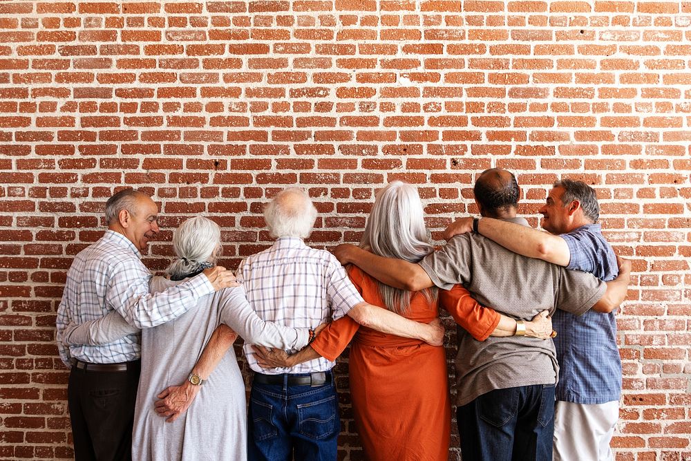 Group of senior people standing together, arms around each other, facing a brick wall. Diverse seniors, friendship, unity…