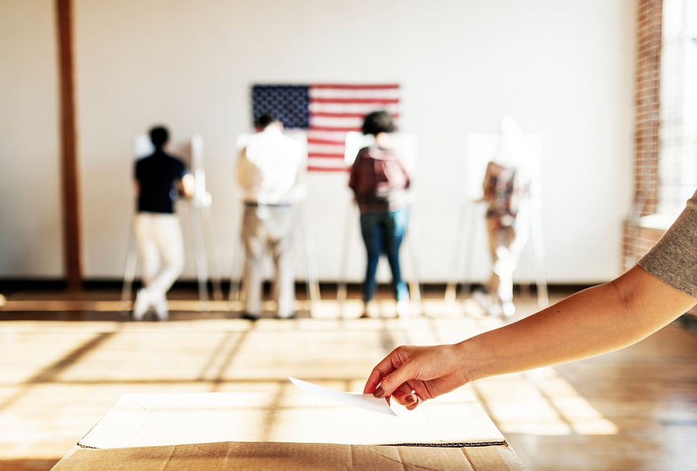 Hand placing a ballot with diverse people voting in a background with an American flag. Voting booths lined up. Hand placing…