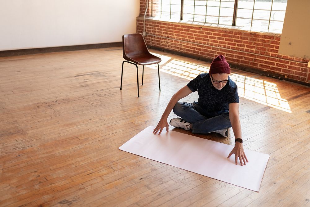 Startup business man sits on wooden floor, arranging a large sheet of paper. Empty room with brick walls and a chair. Focus…