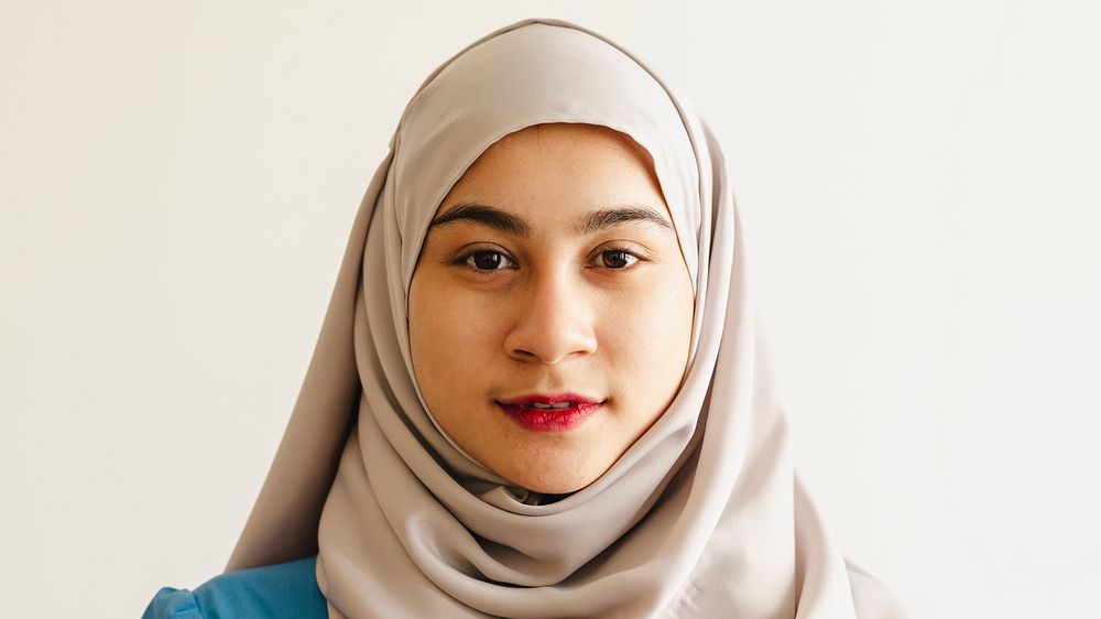 Portrait of a muslim woman wearing a light-colored hijab and blue top, looking directly at the camera with a neutral…