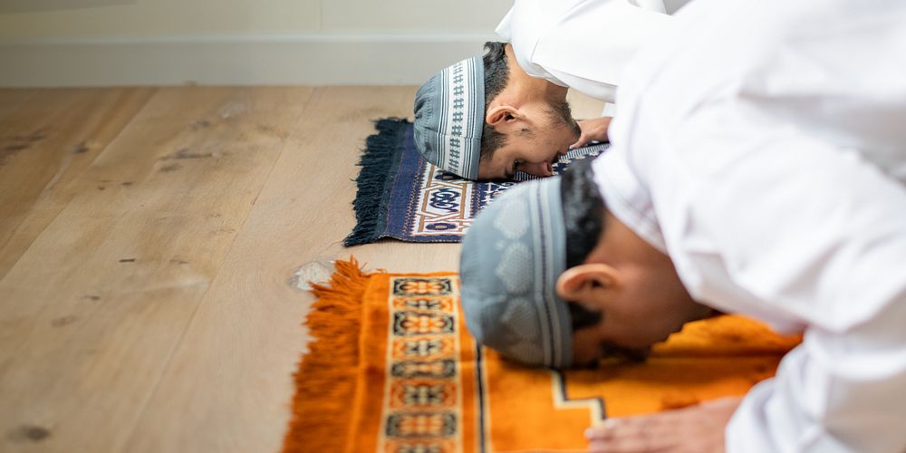 Two muslim men praying on rugs, wearing traditional attire and caps, engaged in prayer. Focus on prayer, traditional attire…