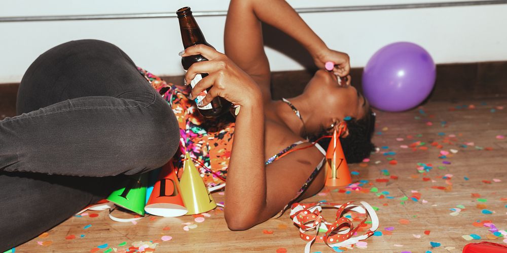 Person celebrating at a party, lying on the floor with a drink, surrounded by confetti and party hats. Festive atmosphere…