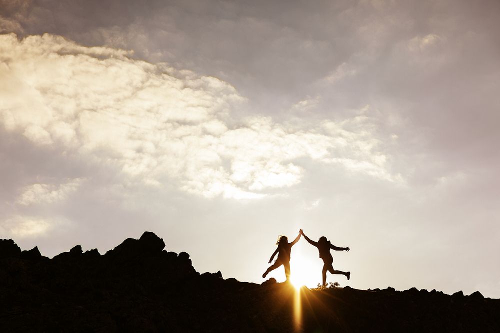 Silhouettes of two people jumping on a rocky hill at sunset. Joyful silhouettes against a dramatic sky. Energetic jump…