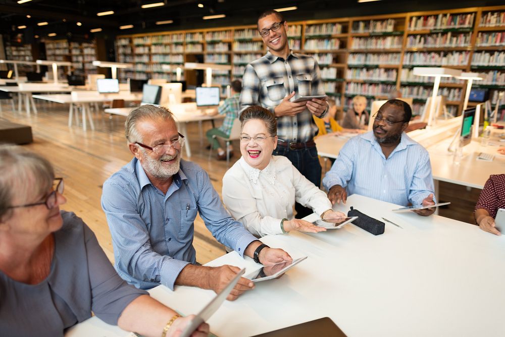 Group of diverse adults in a library, smiling and using tablets. Engaged in discussion, diverse group enjoying technology in…