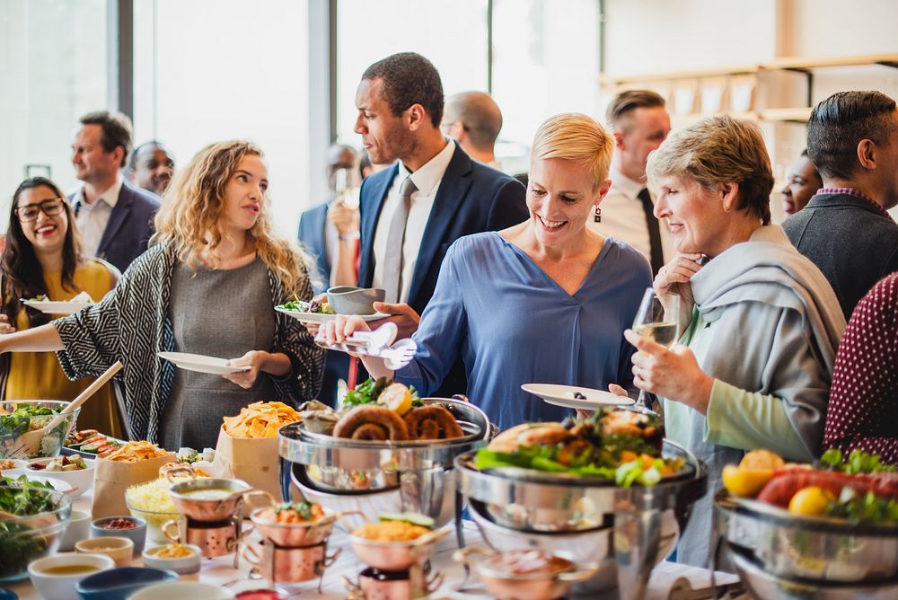 A diverse group of people enjoying a buffet at a social event. Men and women of various ethnicities engage in conversation…