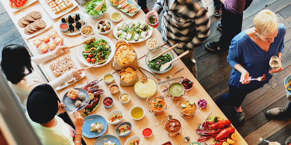 A diverse group of people enjoying a vibrant food spread. The table is filled with colorful dishes, salads, and desserts…