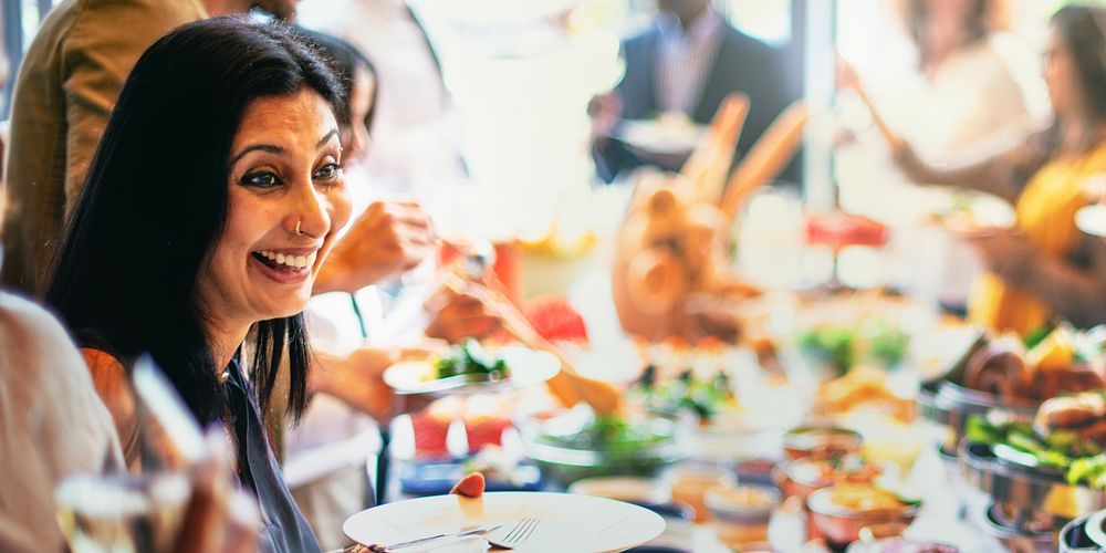 A joyful South Asian woman enjoys a vibrant buffet at a social gathering. The South Asian woman smiles brightly, surrounded…