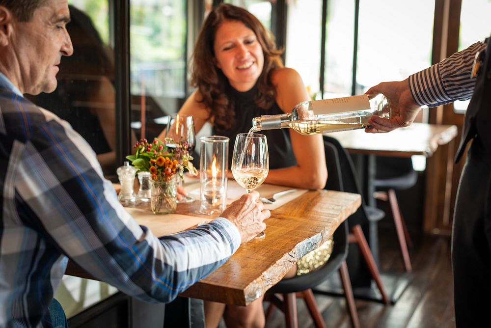 A couple enjoys wine at a restaurant. A waiter pours wine into a glass. The couple smiles, enjoying their meal. Wine…