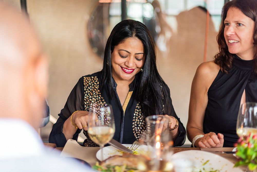 A joyful South Asian woman enjoys a meal at a dinner table, showcasing her vibrant attire. The South Asian woman smiles…