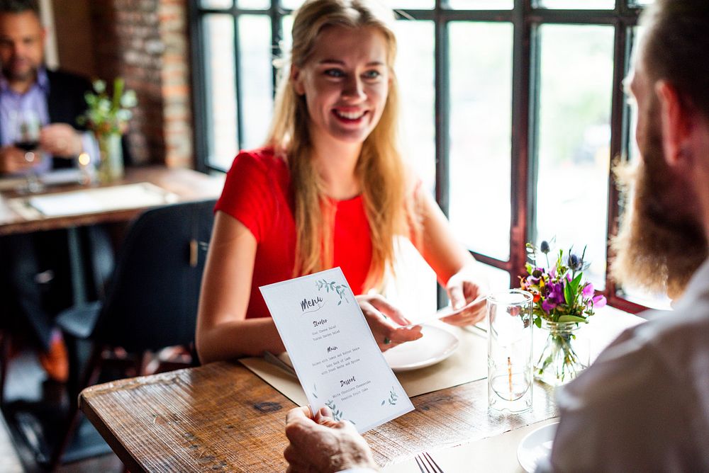 A woman in a red shirt smiles at a man in a cozy restaurant. Bright atmosphere with flowers on the table and large windows…