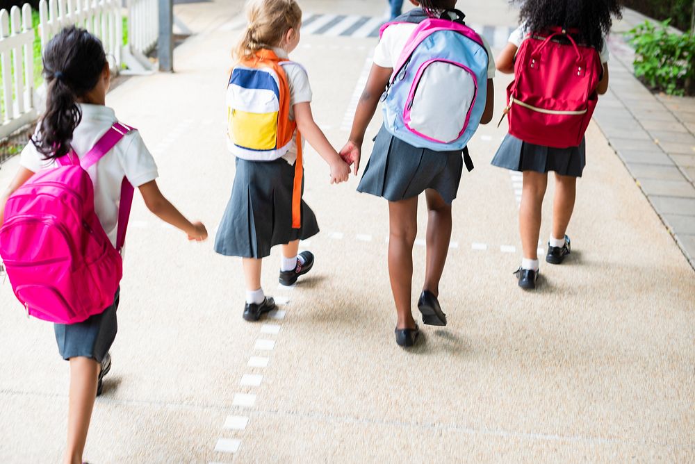 Four young girls with backpacks walk together on a path. School uniforms, backpacks, and friendship are highlighted in this…