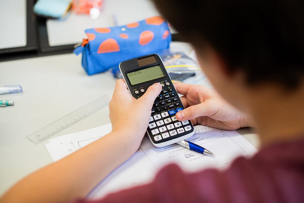 A person using a calculator at a desk with a pen and papers. The calculator is a key focus, with a blue pencil case nearby.…