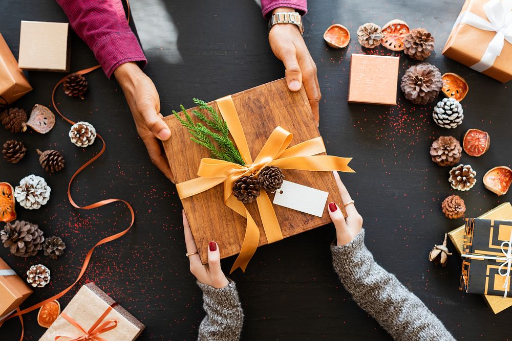 Hands exchanging a gift box with a ribbon. Gift exchange, holiday theme. Pinecones and dried oranges surround the gift.…