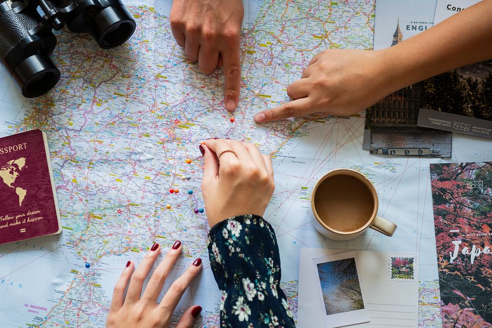 Hands pointing at a map with travel items like a passport, coffee, and photos. Planning a trip with a map, travel…