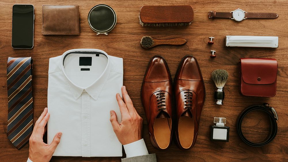 Neatly arranged men's accessories: white shirt, brown shoes, tie, wallet, watch, and grooming items on a wooden surface.…