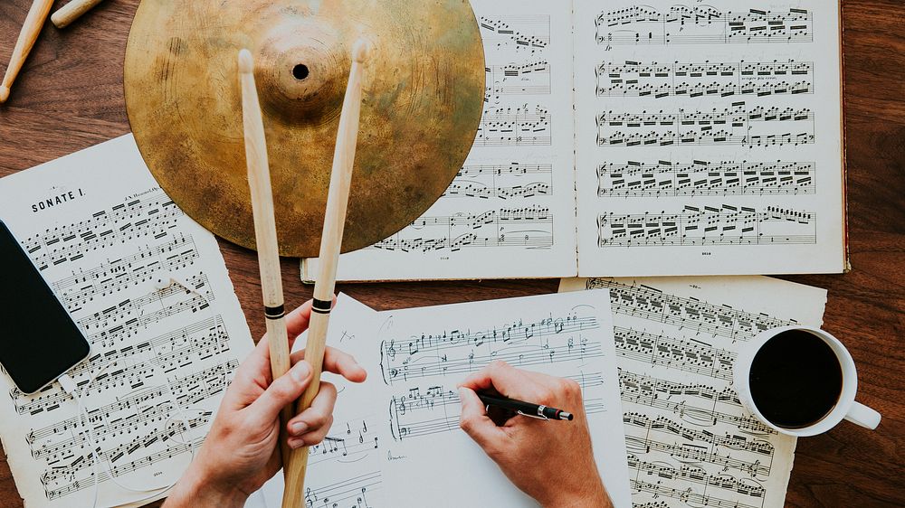 Hands holding drumsticks over sheet music, writing notes. Musical composition, sheet music, and drumsticks on a wooden…