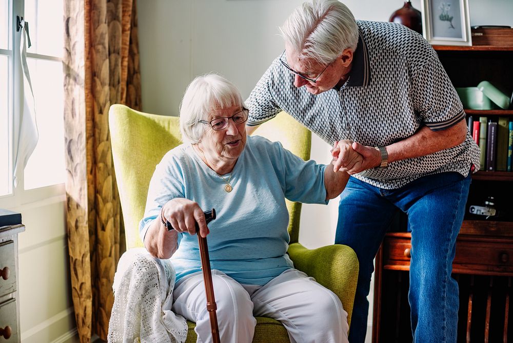 Elderly couple in a cozy room. An elderly man helps an elderly woman with a cane. The elderly couple shares a moment in a…