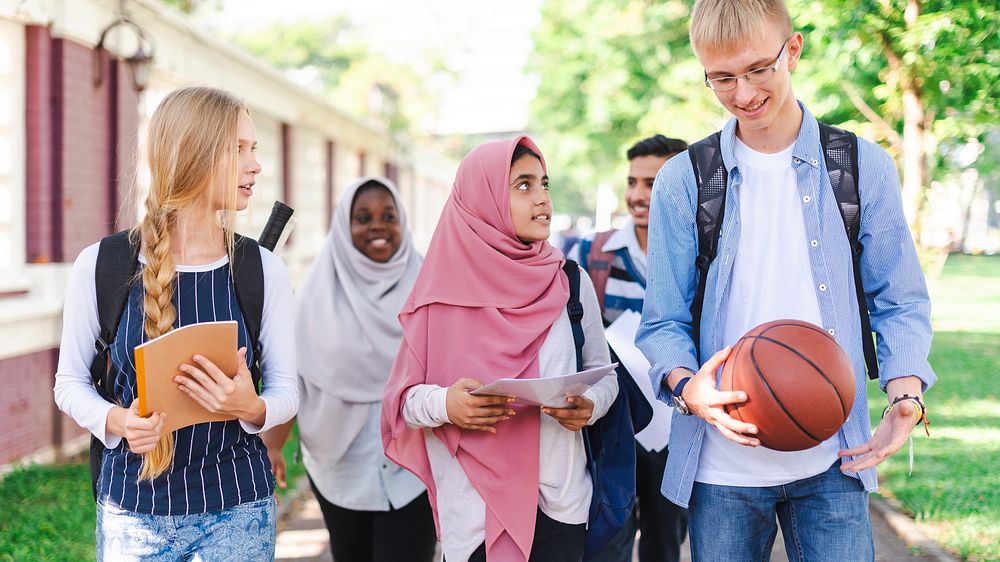 Diverse group of students walking outdoors, carrying books and a basketball. They are smiling and engaged in conversation…