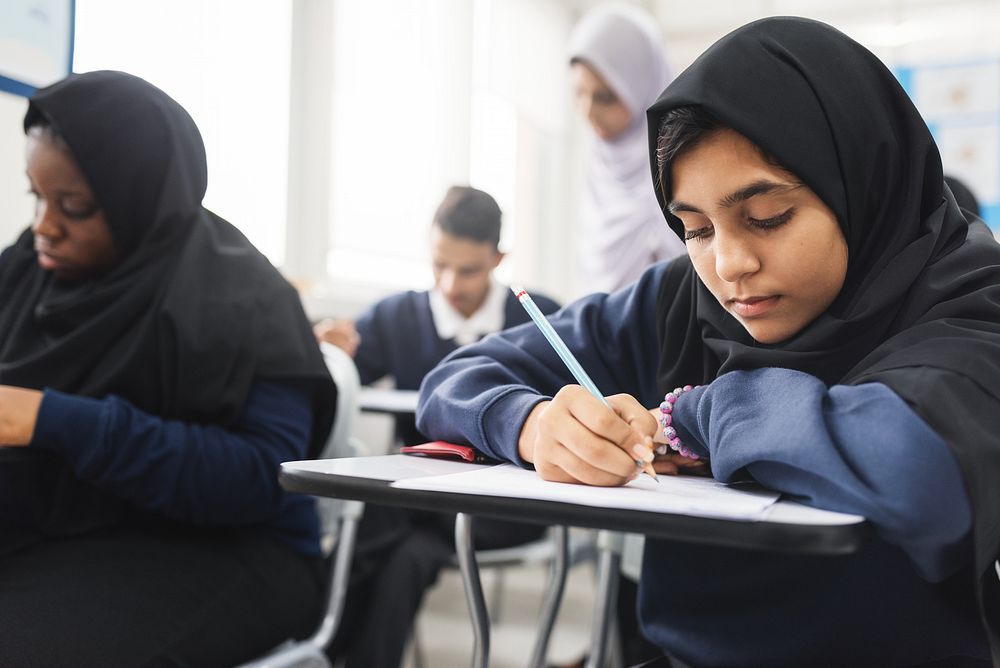 Muslim girl and students focused on writing in a classroom. Diverse group of students, including a girl in a hijab, engaged…